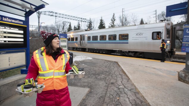 Une femme portant un manteau et un dossard distribue des cafés à l'extérieur d'une gare.