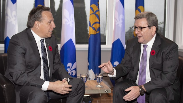 Le premier ministre François Legault (à gauche) discute avec le maire de Québec, Régis Labeaume (à droite), lors d’une rencontre à la salle Mistrale de l’Aquarium du Québec le 1er novembre 2018. Les deux hommes sont assis dans des fauteuils en cuir. Derrière eux, on aperçoit des drapeaux du Québec et de la Ville de Québec.
