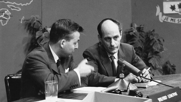Dans un studio de télévision, assis à une table surmontée d'un micro de table, l'animateur Wilfrid Lemoine discute avec René Lévesque, nouveau député du Parti libéral du Québec, dans la circonscription de Montréal-Laurier.