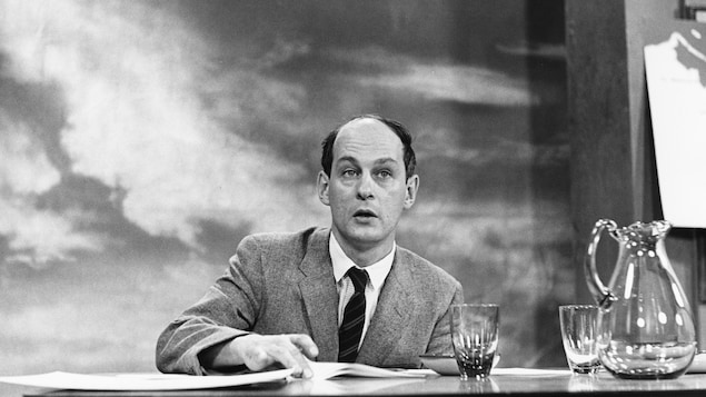 Dans un studio de télévision, assis à une table où reposent une carafe et deux verres, l'animateur René Lévesque s'apprête à tourner avec son index la feuille devant lui.