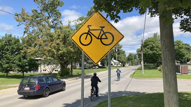 Des cyclistes sur une route de l'île d'Orléans avec un panneau jaune comprenant un vélo. Une auto les contourne par la gauche.
