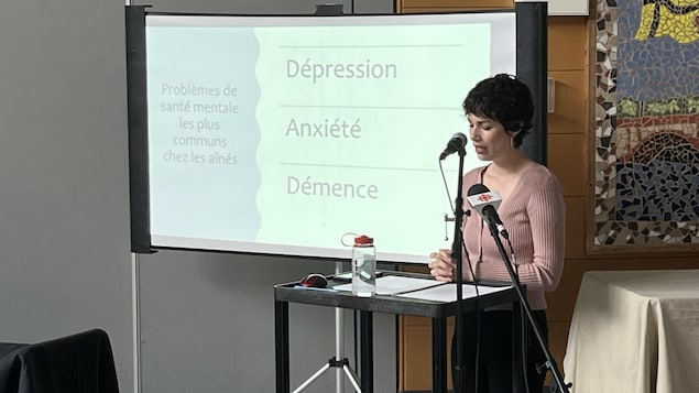 Justine Aubut-Beaudry parle, debout derrière une table, u ne projection powerpoint affichée derrière elle,lors de l'AGA du Réseau Santé en français de la Saskatchewan, à Saskatoon, le 28 mai 2022.