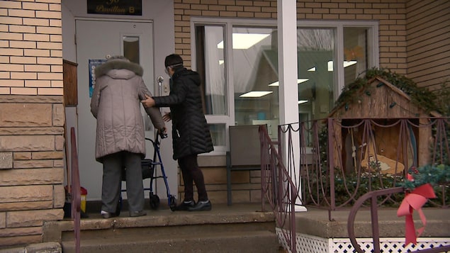 Deux femmes, de dos, entrent dans une résidence. On voit des décorations de Noël à l'extérieur.