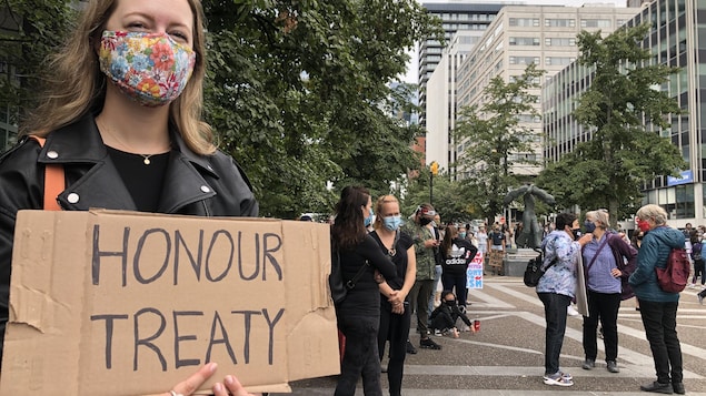 Une manifestante portant un couvre-visage tient une pancarte en carton sur laquelle on peut lire « Respectez les traités ».