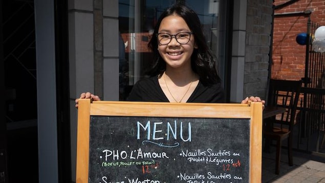 Une adolescente souriante montre le menu de l'établissement, debout devant le restaurant situé à Gatineau.