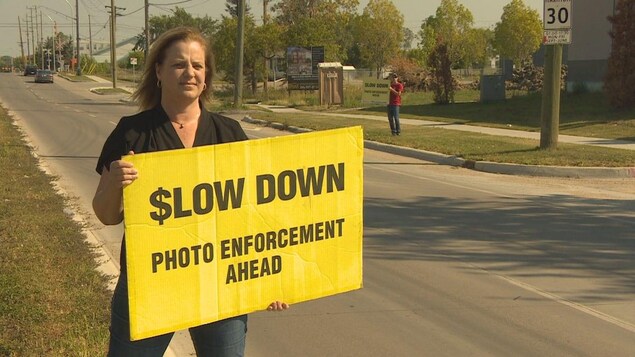 Une femme qui tient une affiche jaune sur laquelle on lit (en anglais) : ralentissez, photo radar devant. En arrière-plan, on voit la rue et un autre homme avec un affiche semblable. Une femme qui tient une affiche jaune sur laquelle on lit (en anglais) : ralentissez, photo radar devant. En arrière-plan, on voit la rue et un autre homme avec un affiche semblable.