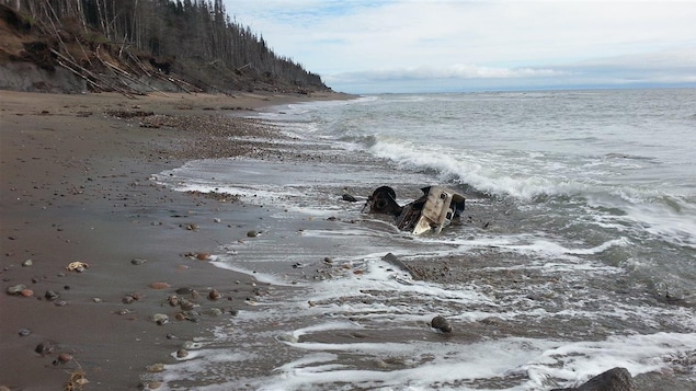 Des débris de ce qui ressemble à un reste de voiture sont enfouis dans le sable au travers des vagues du Saint-Laurent