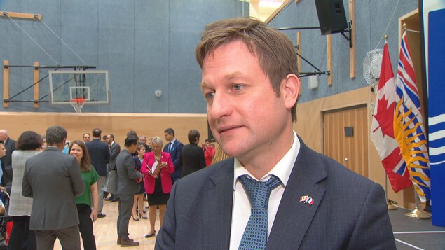Plan serré de Rob Fleming en entrevue dans un gymnase d'école avec des gens qui discutent en arrière-plan. Plan serré de Rob Fleming en entrevue dans un gymnase d'école avec des gens qui discutent en arrière-plan.