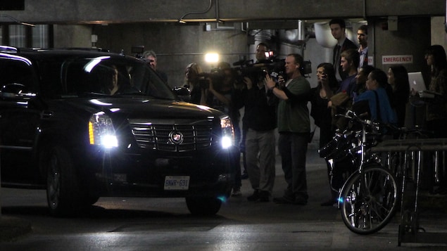 Le maire, au volant de son véhicule, entre dans le stationnement souterrain devant une foule de journalistes qui l'attendent.