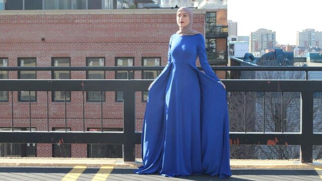 Un mannequin porte une robe bleue et un voile beige. Elle pose sur une terrasse.