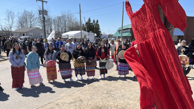 Un vestido rojo cuelga de un árbol y flota en el viento. Detrás, una docena de mujeres forman una fila con un tambor tradicional en la mano.