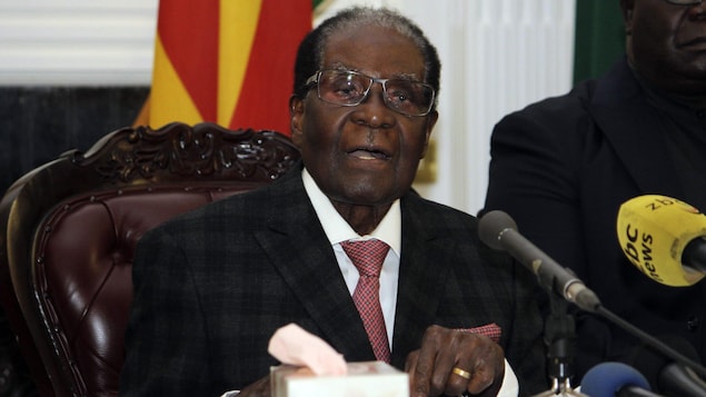 Robert Mugabe est assis sur un fauteuil, derrière un bureau sur lequel se trouvent des micros. Robert Mugabe est assis sur un fauteuil, derrière un bureau sur lequel se trouvent des micros.