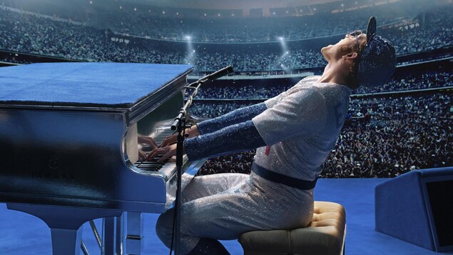 Un homme vêtu d’un uniforme de joueur de baseball en tissus brillant chante et joue du piano.