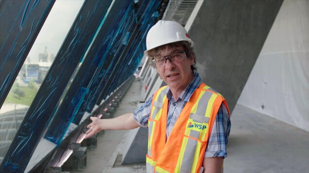 On voit M. Rodrigue, en tenue de travail, parler à la caméra. Derrière lui, on aperçoit la structure de la tour du stade et les nouvelles fenêtres. On voit M. Rodrigue, en tenue de travail, parler à la caméra. Derrière lui, on aperçoit la structure de la tour du stade et les nouvelles fenêtres.