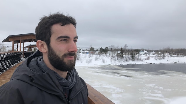 Rodrigue Turgeon, portant un manteau noir, regarde vers la rivière sous un ciel nuageux et sombre. Rodrigue Turgeon, portant un manteau noir, regarde vers la rivière sous un ciel nuageux et sombre.