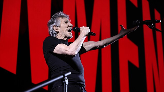 Un homme habillé en noir sur scène tient un micro et chante devant un écran sur lequel apparaissent des lettres rouges sur fond noir. 