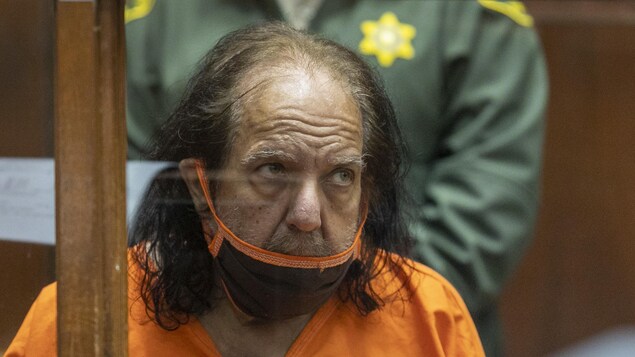 L'homme vêtu d'un habit de prison orange et portant un masque respiratoire se tient assis derrière une paroi vitrée, dans une salle d'audience.