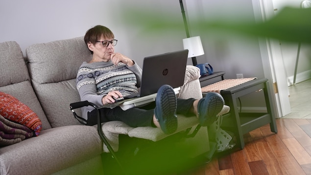 Une dame assise sur un divan travaille avec un ordinateur posé sur ses genoux et avec une cane à ses côtés.