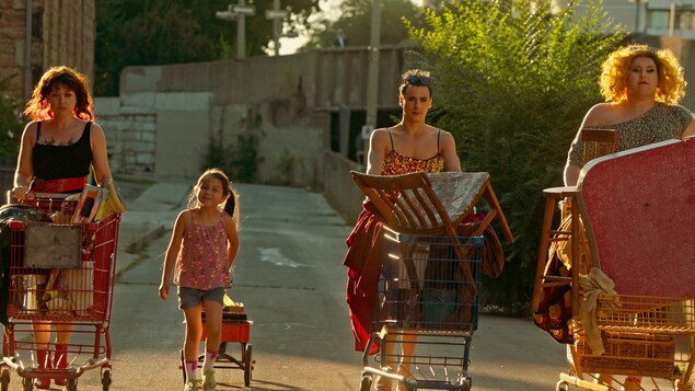 Image d'un film. Dans une ruelle, trois adultes poussent chacun un panier d'épicerie rempli de chaises, tissus et autres objets, alors qu'une petite fille tire un chariot.