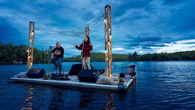 Une chanteuse et un guitariste font une prestation sur un quai flottant au milieu d'un lac, un peu avant le coucher du soleil.