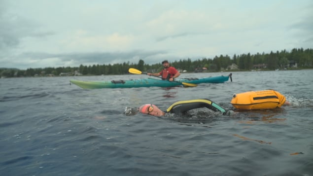 Vêtue de vêtements isothermiques, une personne nage dans les eaux d'un lac. Derrière elle, un homme la suit de près en kayak.