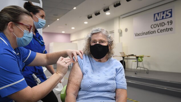 La femme est assise et est entourée d'une infirmière qui la vaccine et d'une femme qui nettoie.
