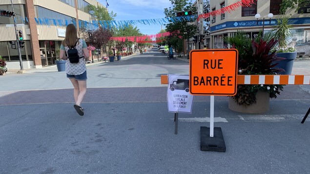 Une pancarte orange indiquant que la rue est barrée aux voitures et une piétonne marche à côté.