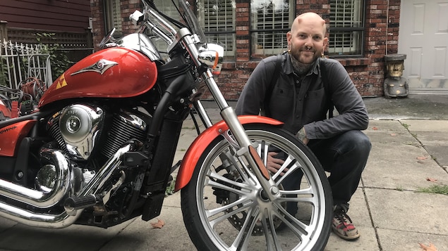 On voit l'homme assis à côté de sa moto orange devant chez lui. On voit l'homme assis à côté de sa moto orange devant chez lui.