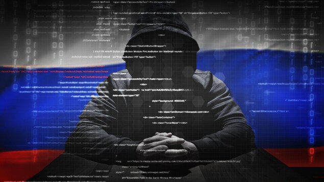 Une photo montrant une personne au visage obscurci par le capuchon de son chandail noir. Derrière lui flotte un drapeau de la Russie.