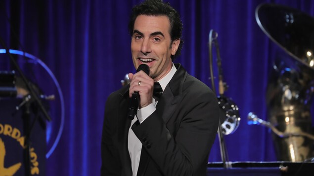L'acteur Sacha Baron Cohen tient un micro sur une scène, entouré d'instruments de musique. L'acteur Sacha Baron Cohen tient un micro sur une scène, entouré d'instruments de musique.