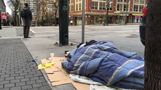 Un homme est enveloppé dans un sac de couchage posé sur des morceaux de carton sur le trottoir et tente de dormir, alors que derrière lui un piéton attend de traverser une intersection.