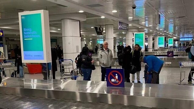 Des passagers attendent leurs bagages à côté d'un carrousel d'aéroport près duquel se trouve un écran publicitaire.