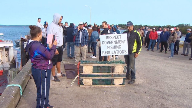 Un homme sur le quai exhibe une affiche sur laquelle est écrit « Respectez les règles gouvernementales sur la pêche ».