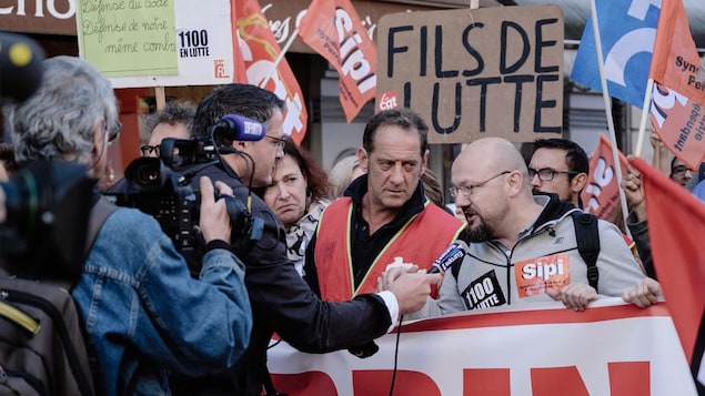 Des hommes tiennent un banderole alors que d'autres brandissent des pancartes derrière eux. Un homme répond au micro d'un journaliste. Des hommes tiennent un banderole alors que d'autres brandissent des pancartes derrière eux. Un homme répond au micro d'un journaliste.