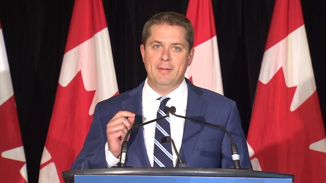 Andrew Scheer, la main levée, regarde la caméra lors d'une conférence de presse.