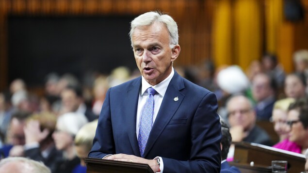 Un député vêtu d'un complet et d'une cravate bleus, debout aux Communes, les mains sur un lutrin, en train de parler.