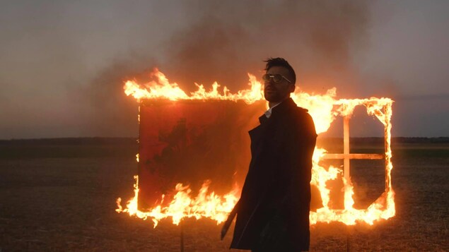 L'artiste pose dans un endroit désterique devant une toile en feu. 
