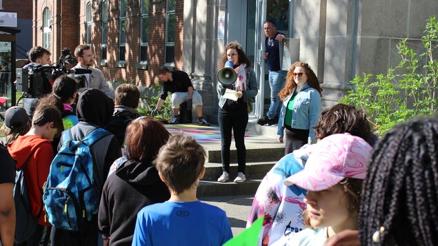 Une dame s'adresse à une foule réunie devant une école primaire à l'aide d'un porte-voix.