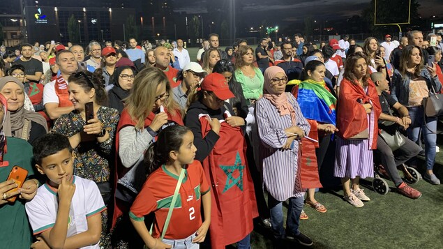 حشد من الناس واقفون ويتقدمهم أشخاص يرتدون علم المغرب.