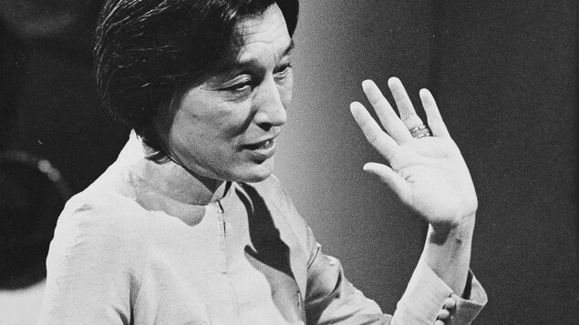 Dans un studio de télévision, l'écrivaine Han Suyin, levant le bras lors d'une entrevue. Un micro de table est posé devant elle.
