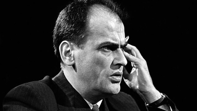 Jean-Paul Desbiens est assis à une table devant un micro et une main tenant une cigarette est appuyée contre sa tempe.