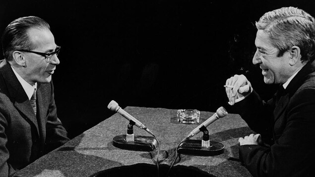 Dans un studio de télévision, Léon Dion s'entretient avec l'animateur Fernand Seguin. Deux microphones sont posés sur le pupitre devant eux.