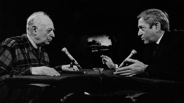 Dans un studio de télévision, le photographe Brassaï discute avec l'animateur Fernand Seguin. Deux microphones sont posés sur le pupitre devant eux.