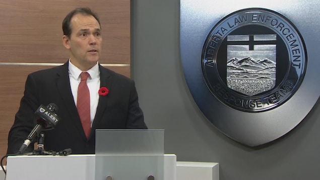 Un homme derrière un podium avec un micro tient une conférence de presse. Il porte un coquelicot accroché sur sa veste. Sur le mur à côé de lui se trouve le logo de l'unité spéciale de police qui se nomme ALERT. Un homme derrière un podium avec un micro tient une conférence de presse. Il porte un coquelicot accroché sur sa veste. Sur le mur à côé de lui se trouve le logo de l'unité spéciale de police qui se nomme ALERT.