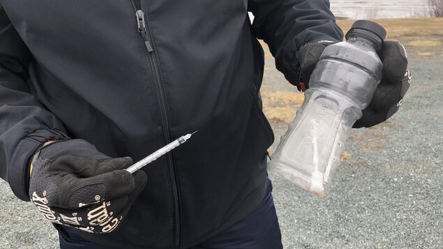 Un homme tient dans sa main droite une seringue usagée , et dans sa main gauche une bouteille de Powerade contenant une deuxième seringue usagé.