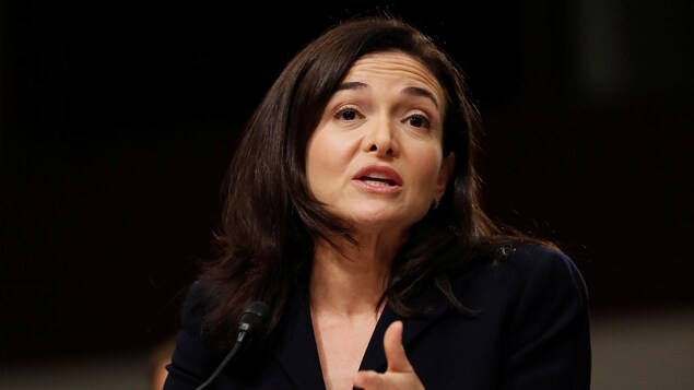 Une photo de Sheryl Sandberg, en tailleur noir, en train de parler devant un micro.
