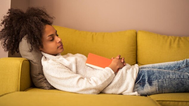 Une jeune femme endormie allongée sur un canapé, un livre ouvert posé sur elle. 