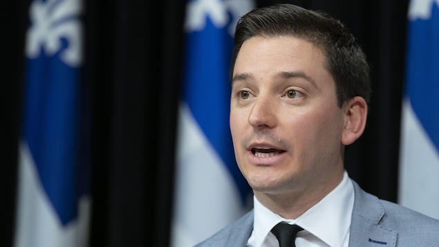 Le ministre Simon Jolin-Barrette parle dans un micro devant une série de drapeaux du Québec. 