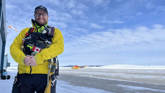 A man stands on a snowy runway with a helicopter behind him. 