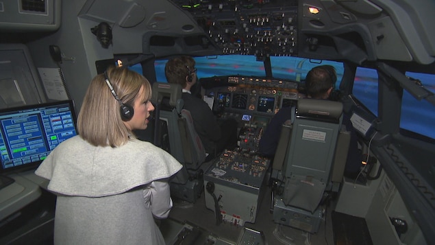 On voit l'intérieur de la cabine du simulateur de vol. Un pilote et un co-pilote sont assis aux commandes (on les voit de dos). Derrière eux, l'instructrice parle.